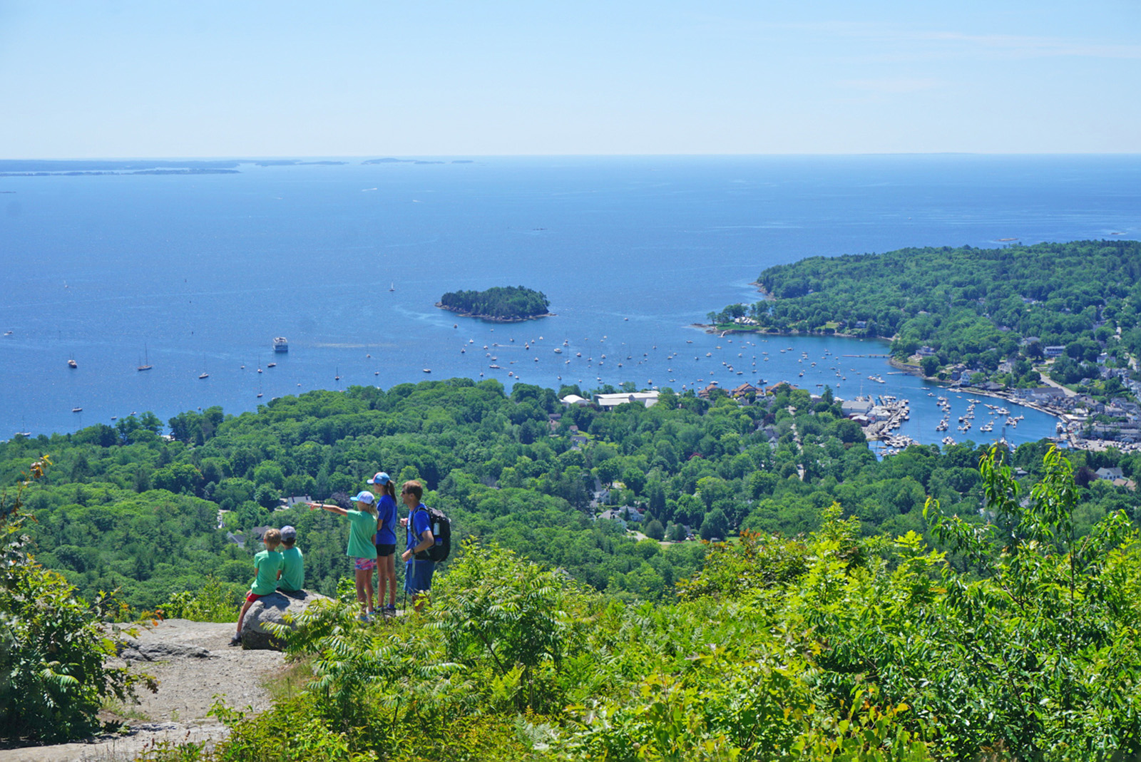 Wide shot of a coastal vista, sailboats visible in water, group of young guests in foreground.