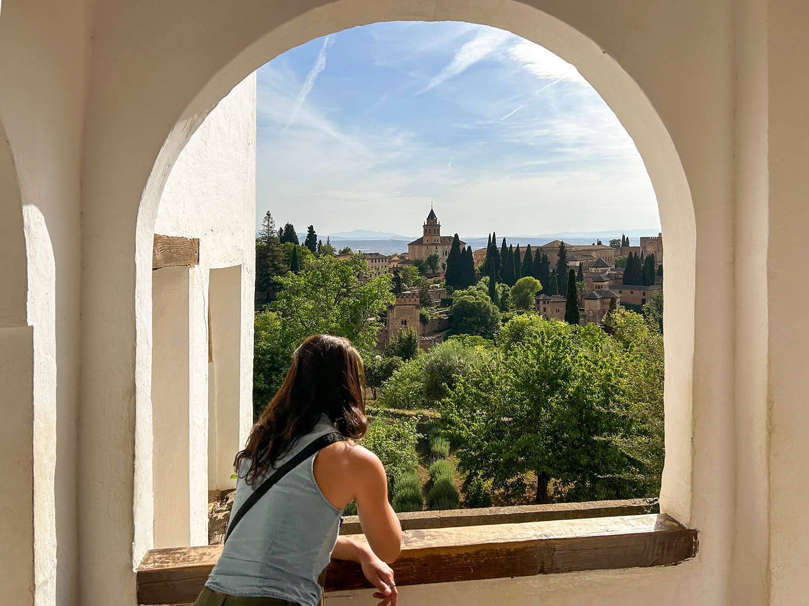 Woman standing next to a large window, looking out to a town surrounded by tall trees