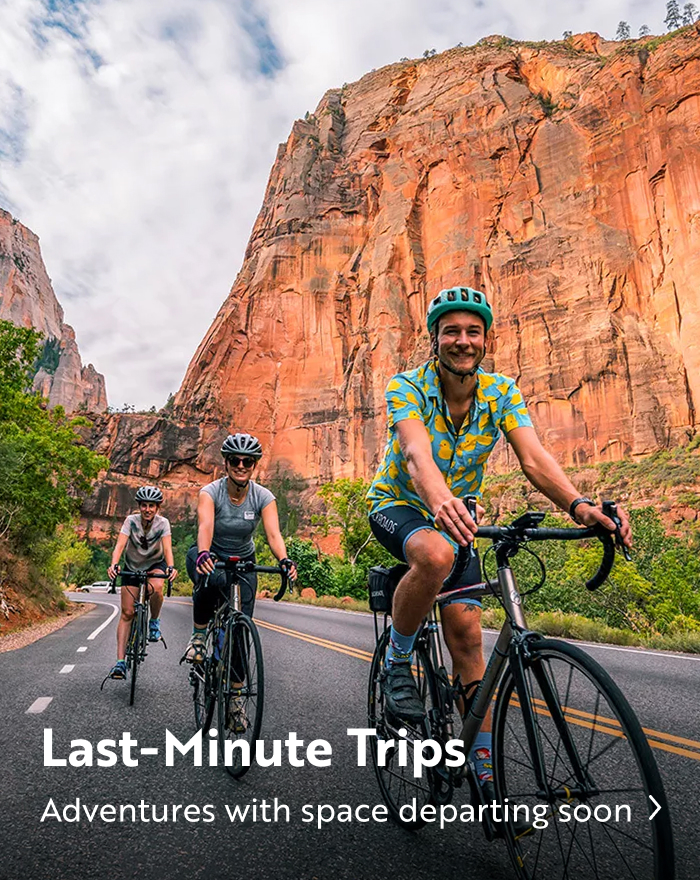 A group of people road cycling on a paved road beneath towering red rock cliffs. Text overlay reads: "Last Minute Trips - Adventures with space departing soon".