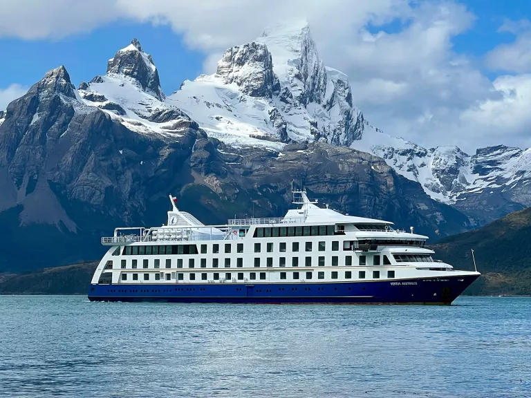 Wide shot of cruise ship, craggy mountains, clouds.