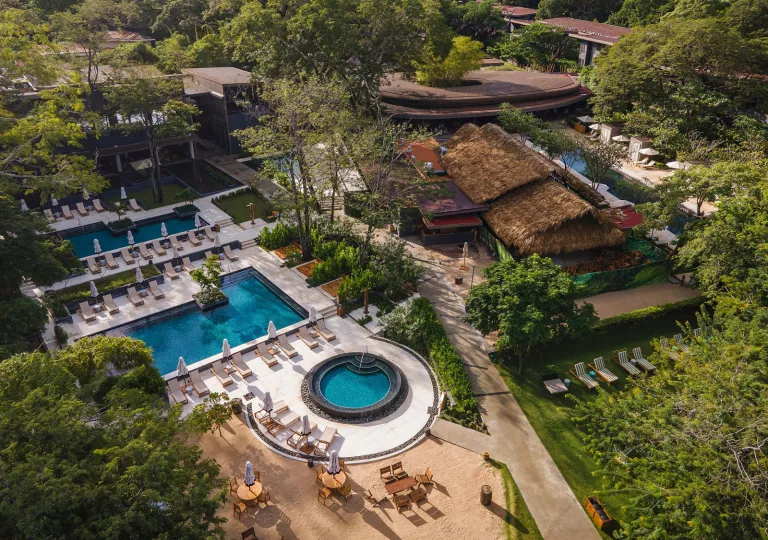 Sky view of outdoor pools and huts, with a modern building in the back