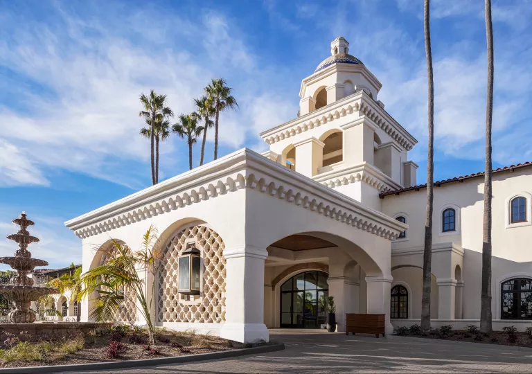 Exterior view of white building with a stone fountain and palm trees in front