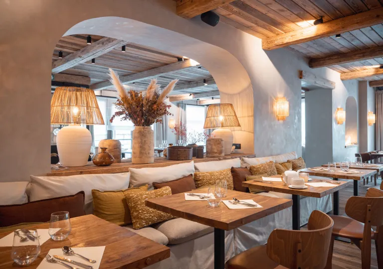 Elegant hotel restaurant with wood-beam ceilings, natural wood furnishings, woven pendant lamps, and cushioned banquette seating. Decorative vases and warm lighting create a refined yet cozy dining atmosphere.