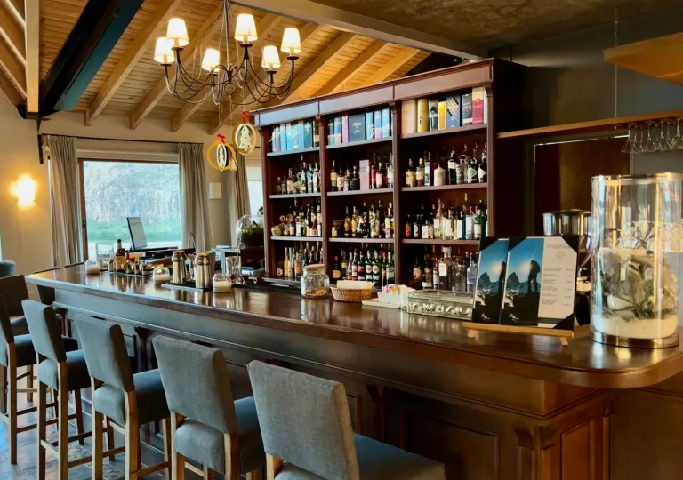 Bar counter with gray stools in front and a wooden shelf with different bottles of alcohol