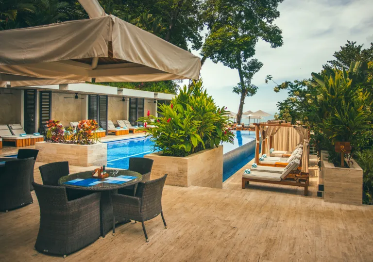 Luxury resort pool area surrounded by tropical landscaping, lounge chairs, and private cabanas. Outdoor dining seating sits beside the pool, with views toward the ocean through the trees in the background.