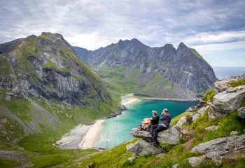 Man and woman on a rocky cliff, looking out to towards larger mountains an a beach