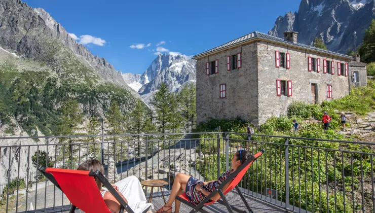 Two women sitting on reclining chairs on a wooden balcony, looking out to large mountains