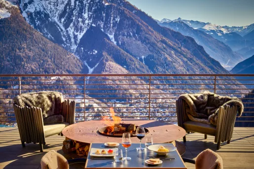 Wooden outdoor patio with wooden chairs around a wood firepit, and large mountains in the background
