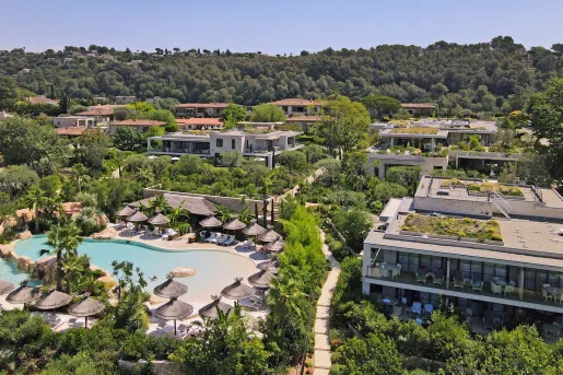 Outdoor pool surrounded by straw umbrellas, next to large hotel villa buildings