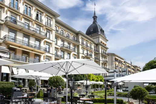 Tables and chairs under large umbrellas, next to a wide hotel building