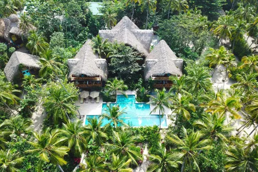 Sky view of hut-style buildings with an outdoor pool in front, surrounded by palm trees