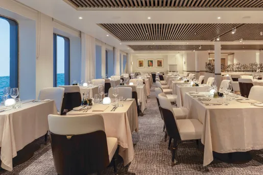 Dining hall with linen over tables and white chairs, with windows to the left looking out towards the ocean