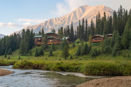 Large grassy valley with two large cabin buildings  looking out to large mountains and a pond
