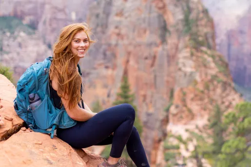 Woman wearing a blue backpack, sitting on an orange boulder with large mountains in the background