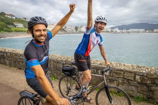 Two men biking on a path with their arms in the air, with a lake in the background