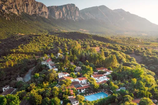 Sky view of a white and red hotel complex in the middle of a forest, with tall mountains in the background