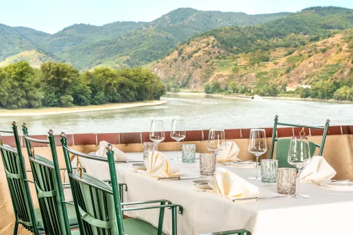 Dining table with green chairs, next to a river surrounded by trees and hills
