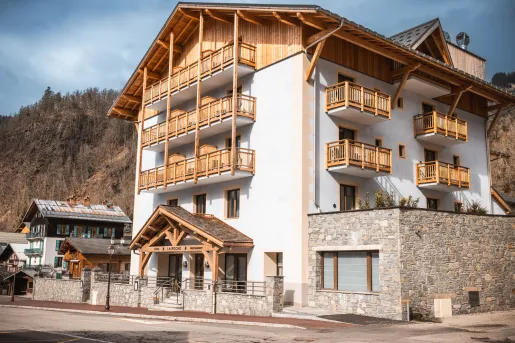 Exterior view of Hotel La Roche, a modern alpine-style hotel with wood balconies, stone accents, and a mountain village setting. The multi-story building is framed by surrounding hills and traditional chalet architecture.