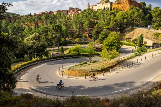 Several cyclists ride around a winding hairpin turn through a forested hillside below a historic village perched on red rock cliffs. Stone buildings and a church overlook the scenic landscape under a partly cloudy sky.