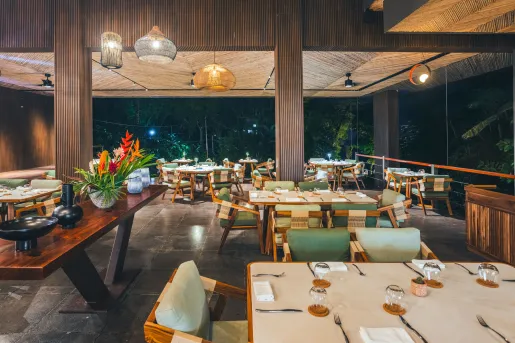 Open-air resort restaurant with wood-paneled architecture, woven pendant lights, and neatly set dining tables. The upscale dining space overlooks lush tropical greenery and is illuminated by warm evening lighting.