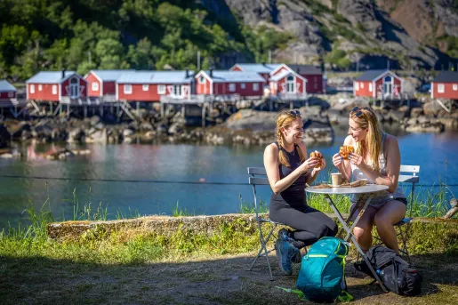 Two women sitting at table eating pastries and drinking coffee in Nusfjord, one of Norway's oldest and best-preserved historical fishing villages, featuring traditional red wooden cabins.