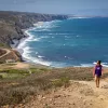 Two hikers walking up a sandy hill with the ocean in the background