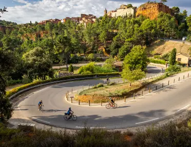 Several cyclists ride around a winding hairpin turn through a forested hillside below a historic village perched on red rock cliffs. Stone buildings and a church overlook the scenic landscape under a partly cloudy sky.