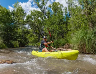 Person paddling a bright green kayak through a shallow river bordered by dense green vegetation.