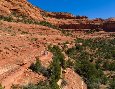Hikers follow a winding trail through a rugged red rock canyon surrounded by desert vegetation and sandstone cliffs.