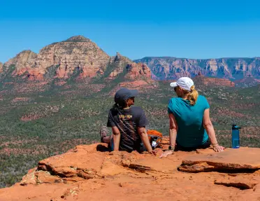 Two travelers sit on a rocky overlook taking in panoramic views of Sedona’s red rock mountains and valleys.
