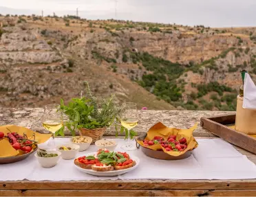 An outdoor table spread features wine glasses, fresh strawberries, bruschetta, and olives set on a white tablecloth.