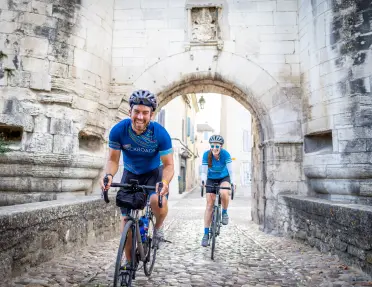 Two cyclists ride on a cobblestone street through a historic stone gateway. Tall medieval walls and an arched passage frame the narrow street in a preserved old town setting.